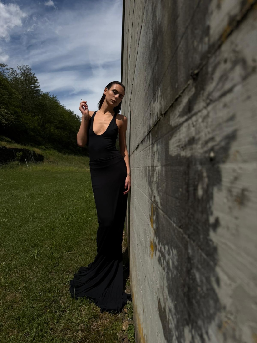 Black gown, leaning on a weathered wall, cigarette, open sky.