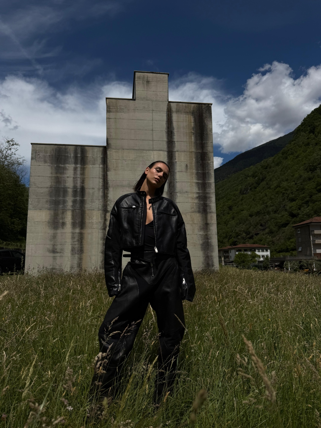 Leather suit before a brutalist silo, alpine landscape.