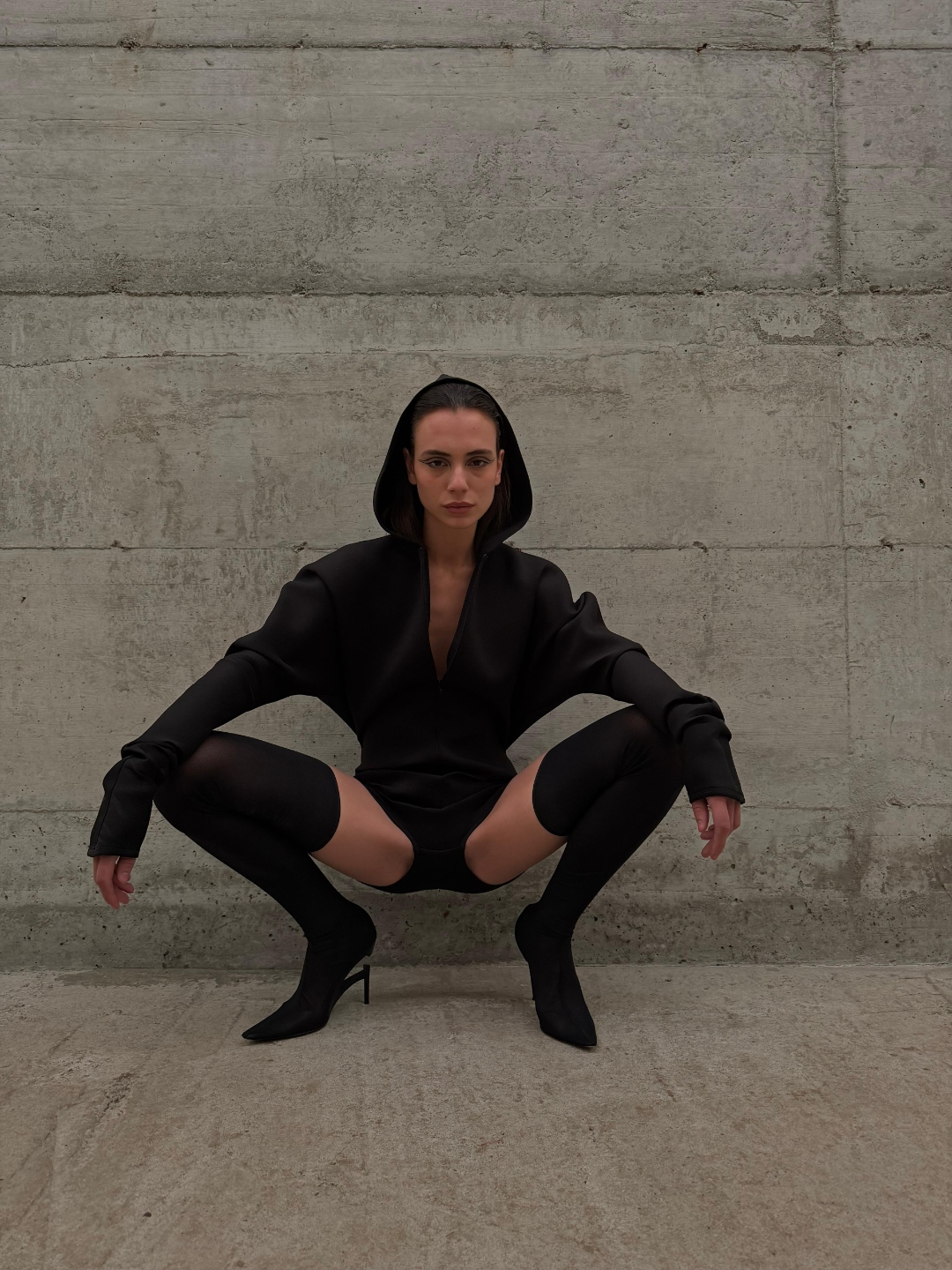 A squatted figure in a hood against a concrete wall.